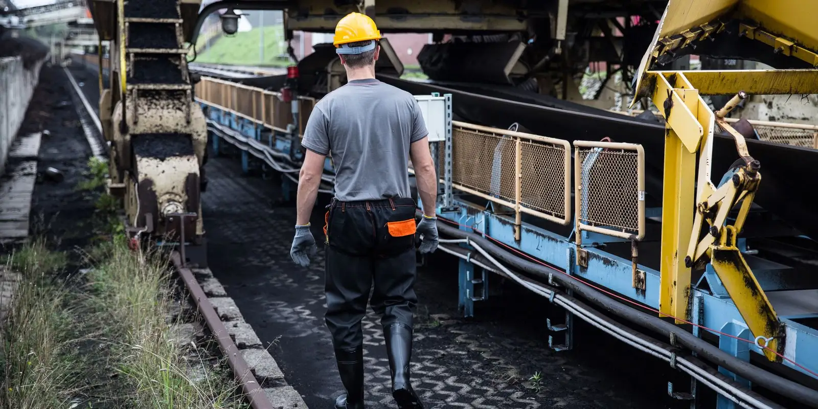 Worker in a hard hat walking an inspection route beside a conveyor at a bulk handling yard